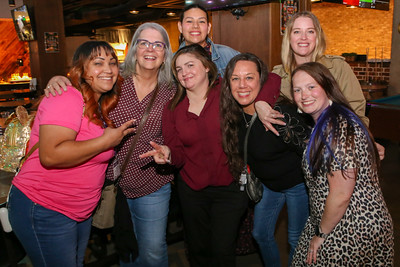Seven women smile and pose together in a lively bar setting, showcasing a mix of casual attire and vibrant expressions.