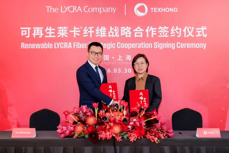 Executives from The LYCRA Company and Texhong International Group shake hands, holding red folders, with a floral arrangement in front.