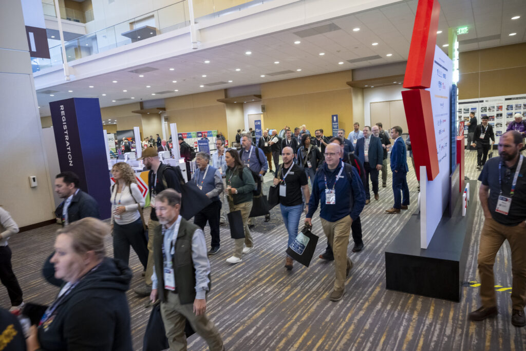 Crowd of attendees walking through a modern convention center, with registration signs and booths visible in the background.