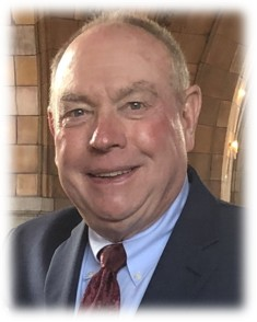 Smiling man in a dark suit with a red tie, set against a softly blurred historic interior background.