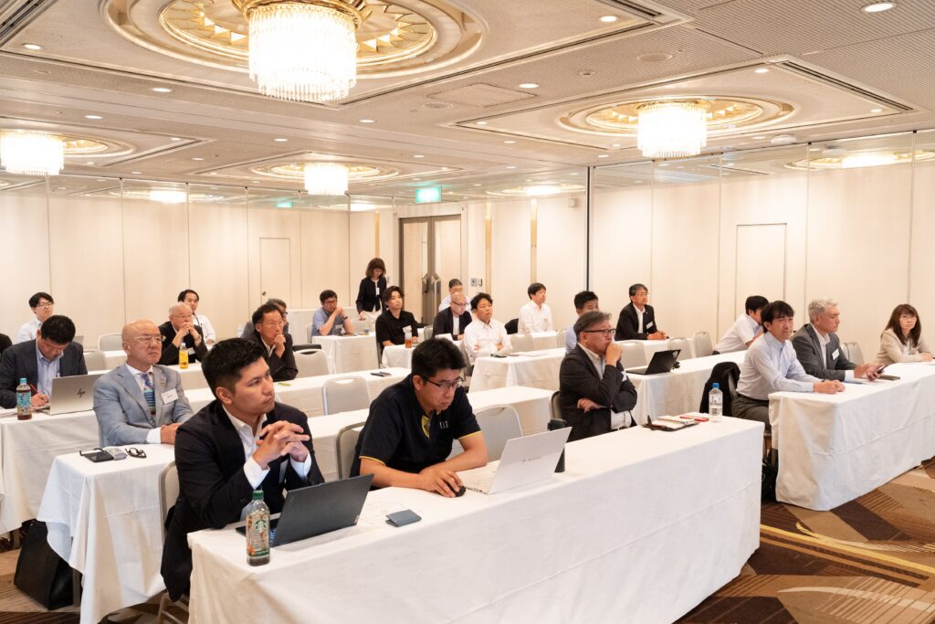 A conference room filled with attendees seated at tables, focused on presentations; some with laptops, soft lighting and modern decor.