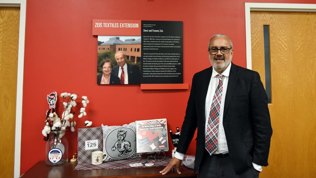 Man in a suit stands next to a display table featuring memorabilia, a photo on the wall, and red backdrop with text.