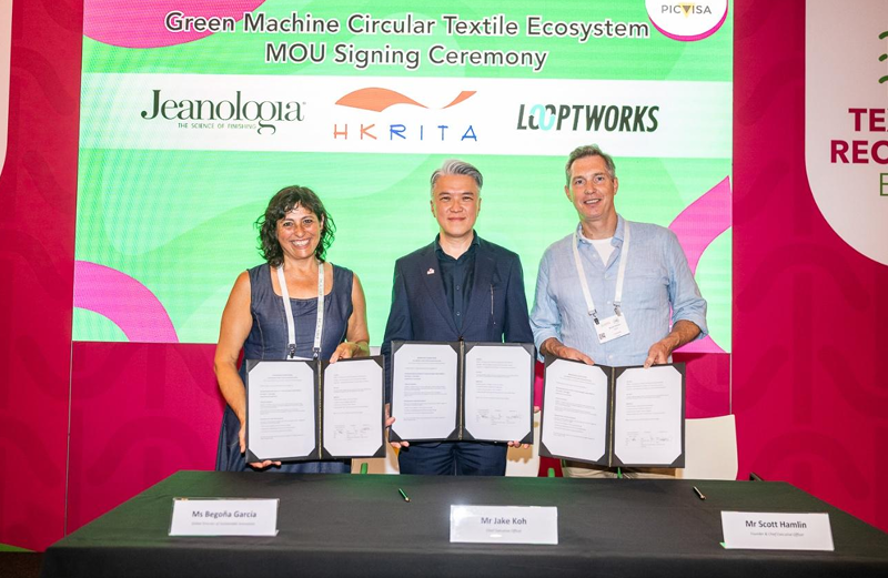 Three individuals stand behind a table holding signed documents, smiling. A colorful backdrop features logos of Jeanologia, HKRITA, and Looptworks.