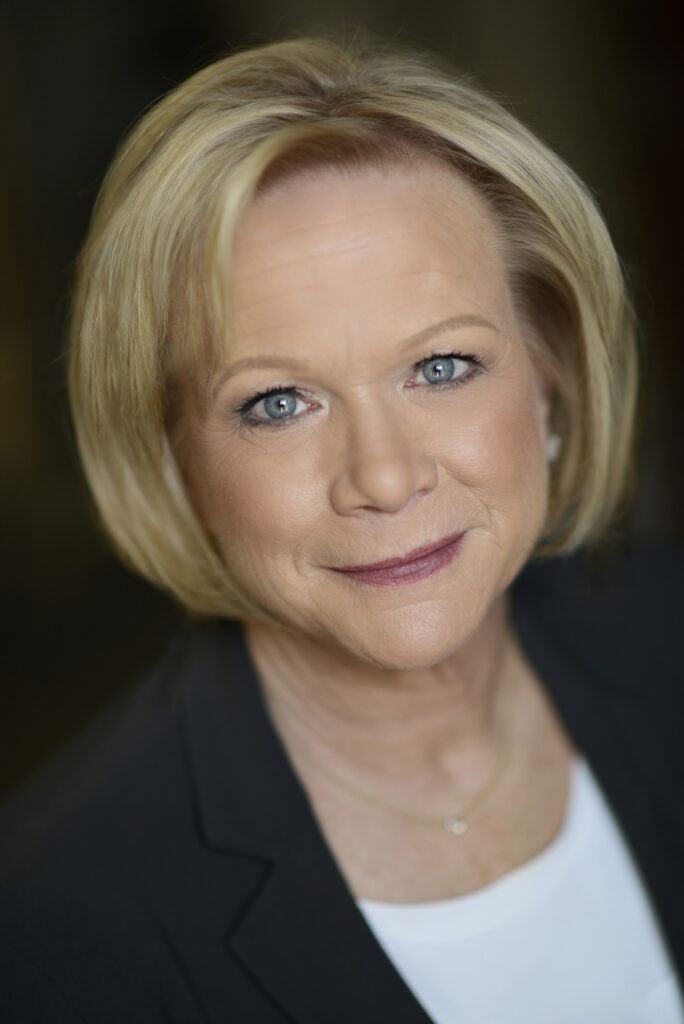 Headshot of Denise Offray, CEO and owner of OTEX Specialty Narrow Fabrics, a woman with blonde bob hair, blue eyes, and a subtle smile, wearing a black blazer over a white top, against a blurred background.