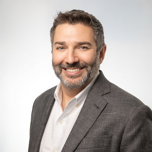 Smiling man with a beard in a blazer over a light shirt, against a softly lit, pale background.