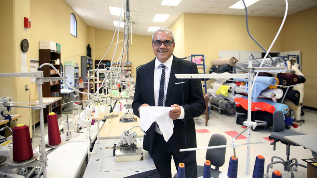 A smiling man in a suit stands in a textile workshop, holding a piece of fabric, surrounded by sewing machines and colorful rolls of material.